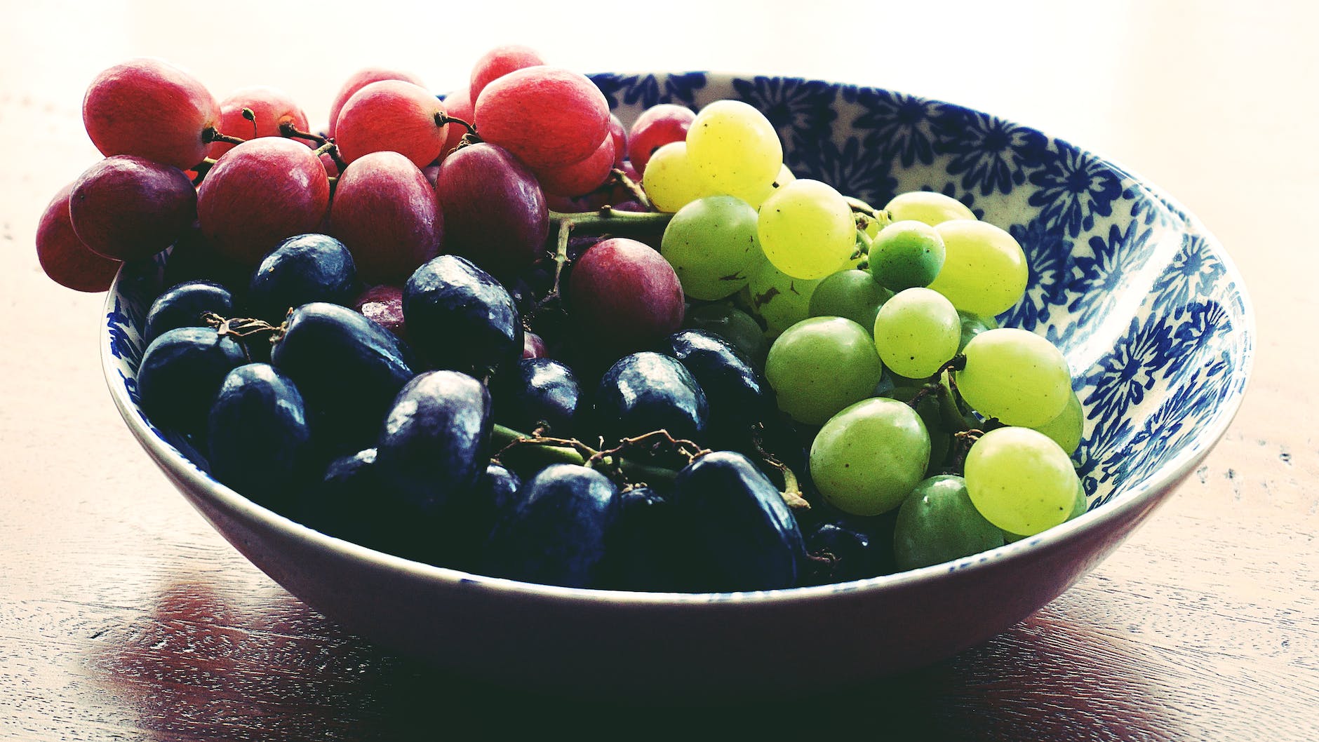 red black and green grapes in round blue and white floral ceramic bowl